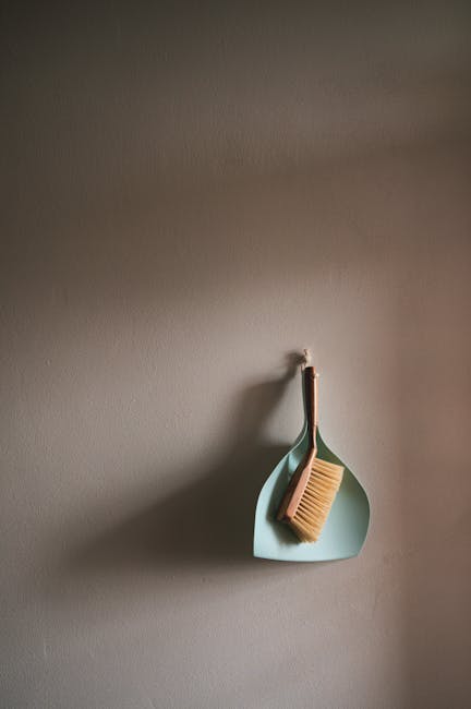 A soft grey wall with a small hook holding a green silicone cleaning brush and a beige wooden scrub brush, neatly arranged on a pale blue oval-shaped wall-mounted container. The tools are clean and ap