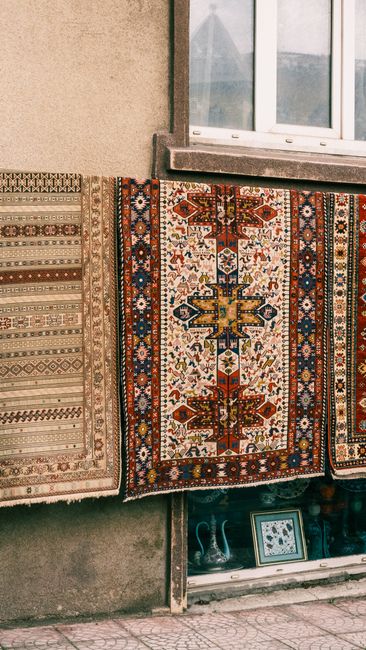 Close-up image showing two traditional oriental rugs with intricate geometric and floral patterns in red, navy, cream, and earthy tones, laid on a rustic white tiled floor. One rug is partially folded